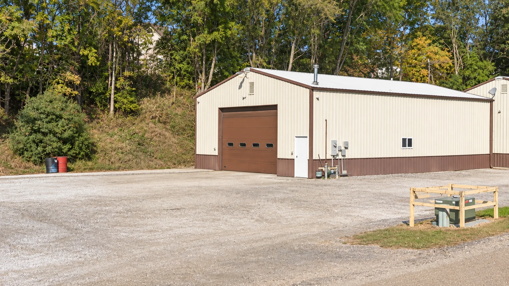Cream metal building with a brown garage door, regular side door, gravel parking lot, and wooded hillside